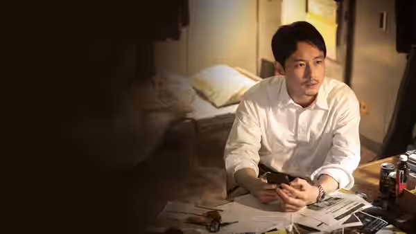 A man in a white shirt sits beside a bed, holding a phone over scattered papers