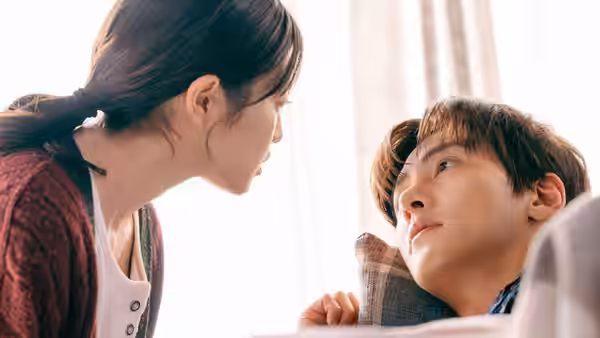 A woman leans close to a man reclining beneath a blanket beside a bright window