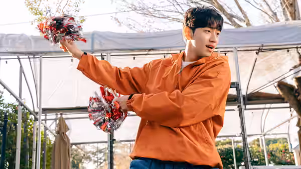 A young man in an orange jacket waves pom-poms beneath an outdoor canopy