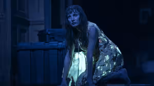 A woman crouches beside rubbish bins in a pale dress and shimmering wrap