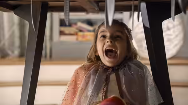 A young girl crouches beneath a table, mouth open beneath dangling knives