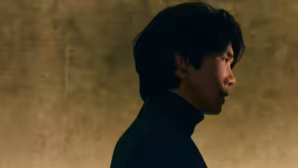 A man in a black turtleneck stands in profile against a bare wall