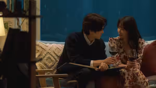 A man and woman sit close together with wine glasses beside a rain-streaked window