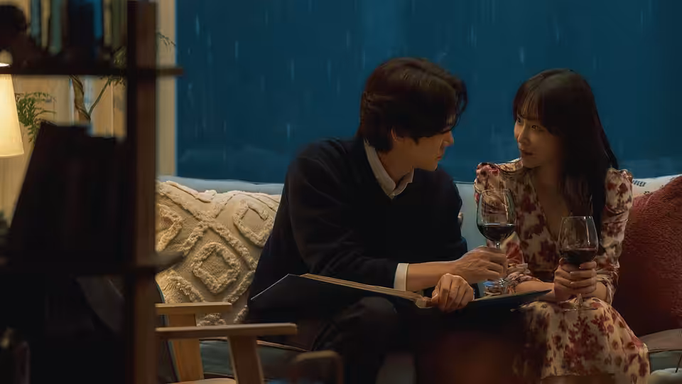 A man and woman sit close together with wine glasses beside a rain-streaked window