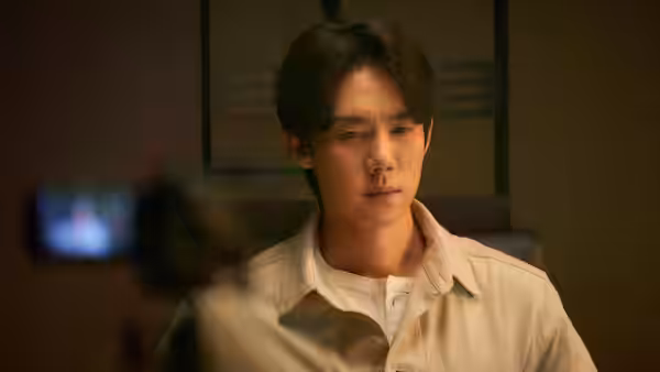 A man in a light shirt faces a camera in a dim room
