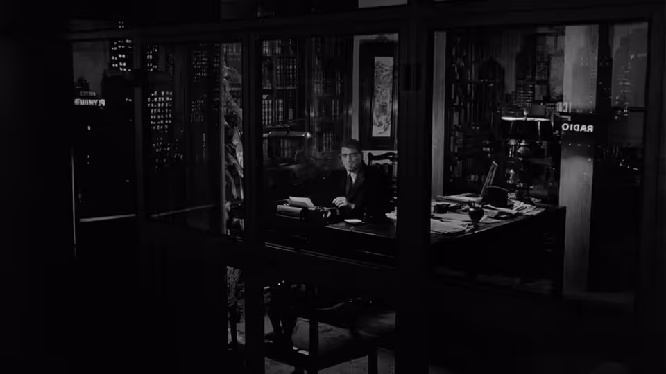 A man sits at a desk in an office, seen through glass partitions