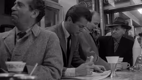 Men gather around a counter in a diner, one writing on a paper