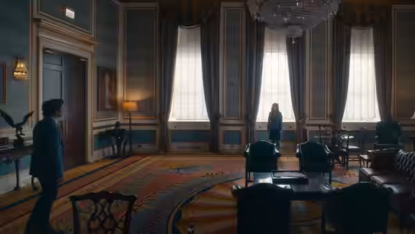 Several people gather in a stately office with tall windows and upholstered chairs