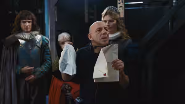 A bald man holds a paper among costumed performers backstage