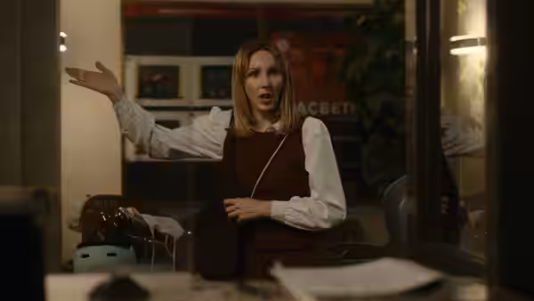 A woman gestures across a backstage room beside a table and bags