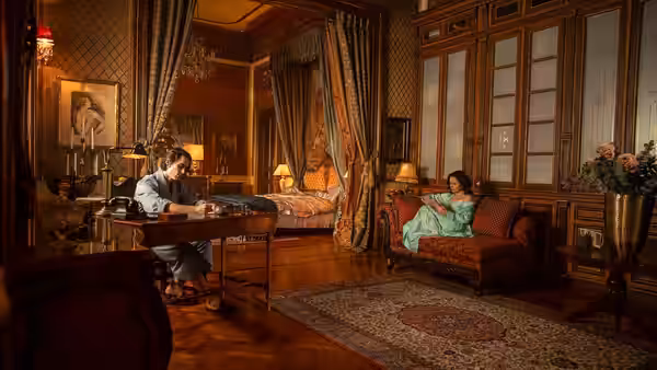 a man works at a desk while a woman sits on a sofa in an ornate room