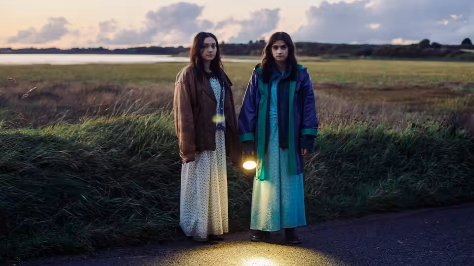 Two young women standing on a rural road at dusk holding a lantern