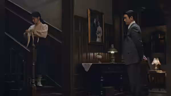A woman pauses on the staircase as a suited man watches from the parlor