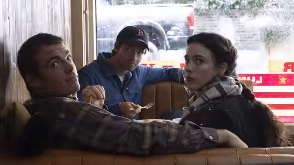 A woman and two men sit in a diner booth, turning toward the camera