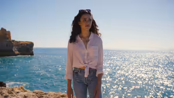 Maya (Margaret Qualley) standing on a rocky shore beside the sunlit sea