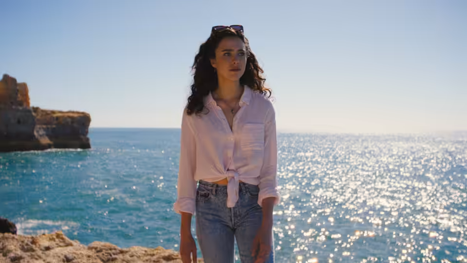Maya (Margaret Qualley) standing on a rocky shore beside the sunlit sea