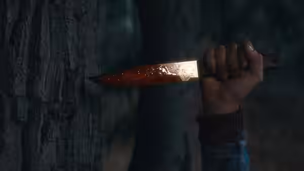 A hand grips a bloodied knife beside a tree in the dark woods