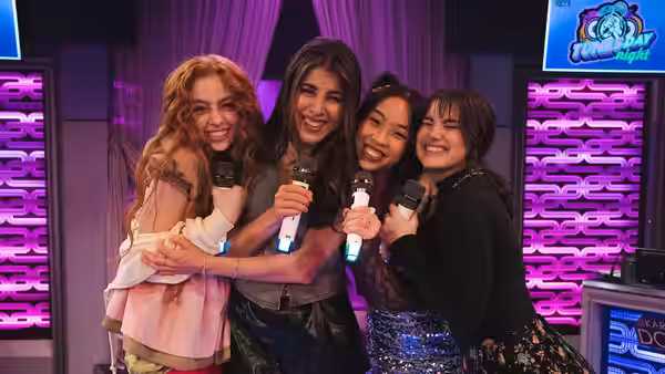 Trina Vega (Daniella Monet) and three women huddle together holding microphones onstage