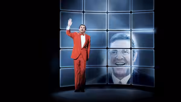 A man in a red suit waving beside a screen showing another man's face