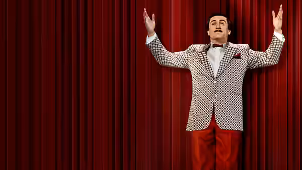 Rupert Pupkin standing with arms raised against red stage curtains