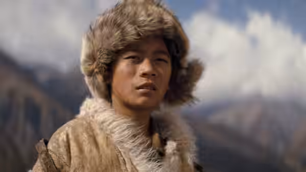 A young man in a fur hat and coat stands before a mountain range