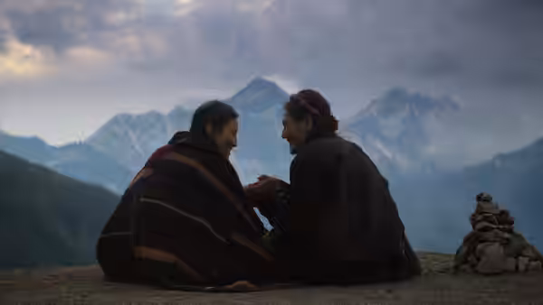 Two men sit wrapped in blankets on a mountain ridge beside a stone cairn