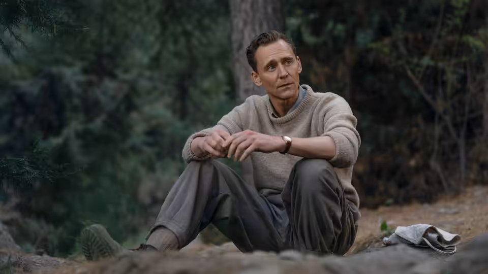 A man in a sweater sits resting on the ground among trees