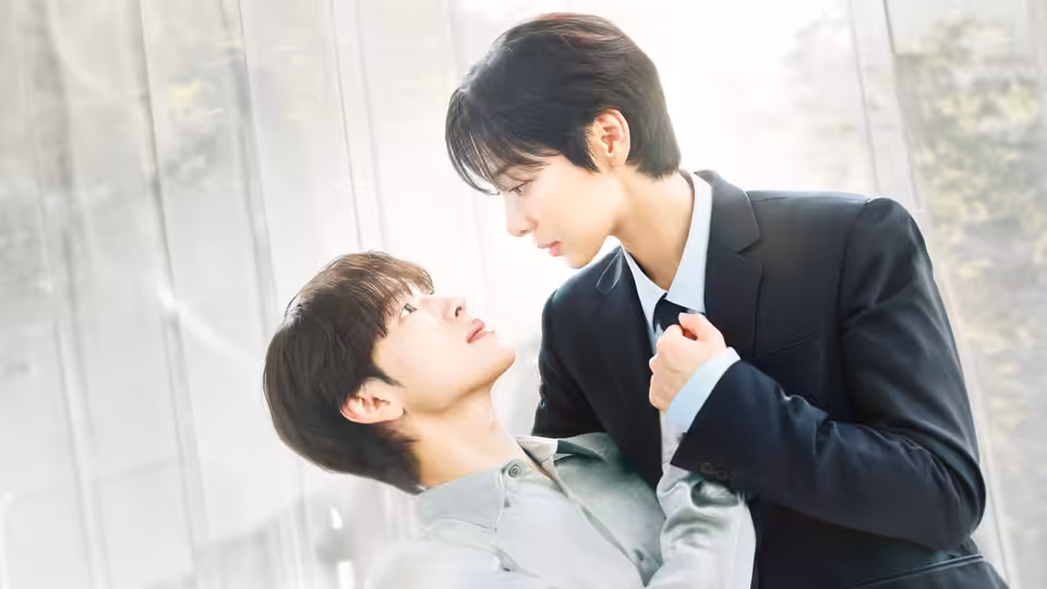A suited man leans over another man reclining against a bright window