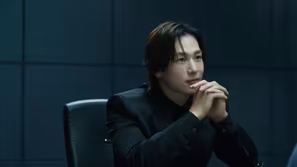 a man in a black suit sitting with hands clasped in a dark room