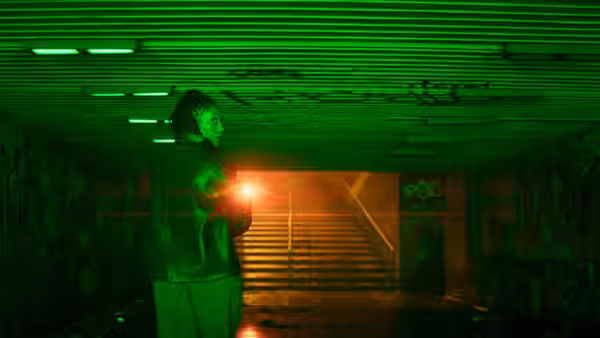 A man holding a glowing light stands beneath a stairway in a graffiti-covered passage