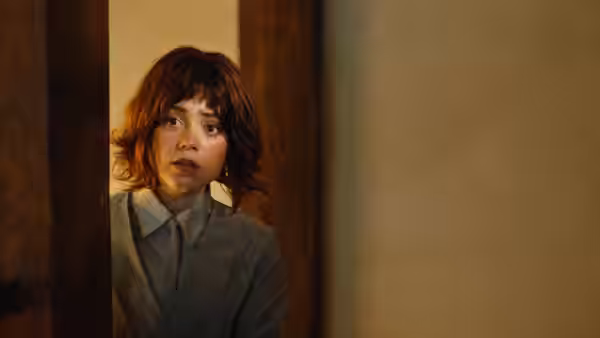 Klara (Jenna Ortega) peers through a doorway in a gray suit