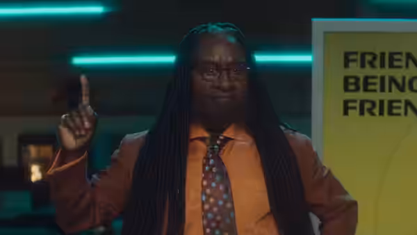 Man with long braids in a suit points upward near a sign