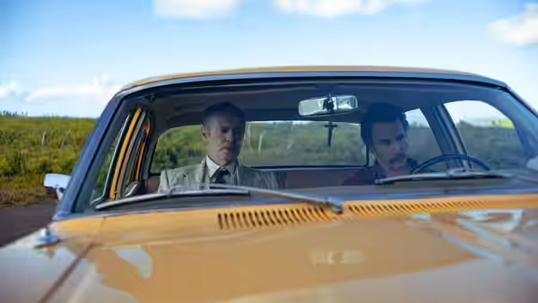 Two men seated inside a yellow vintage car on a rural road
