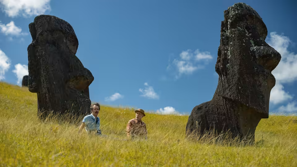 Two men standing on a grassy hillside among large stone Moai statues