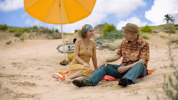 A woman and an older man sitting on the sand beneath a yellow umbrella