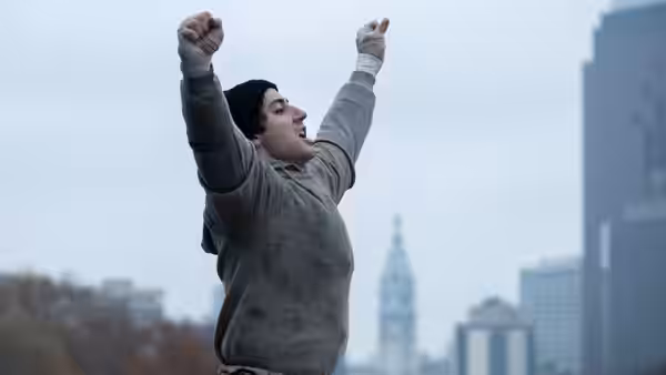 Sylvester Stallone (Anthony Ippolito) raises both arms above a city skyline