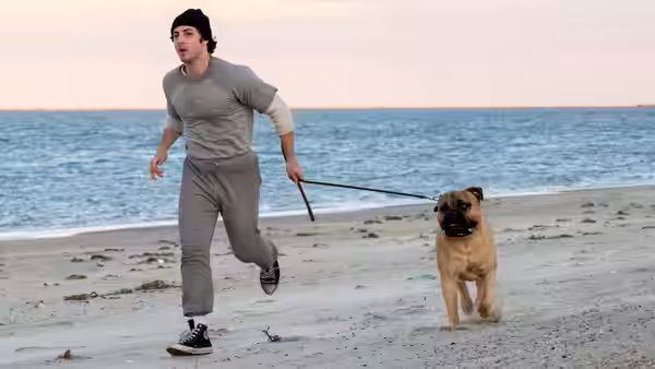 A young man runs on the beach pulling a bulldog on a leash