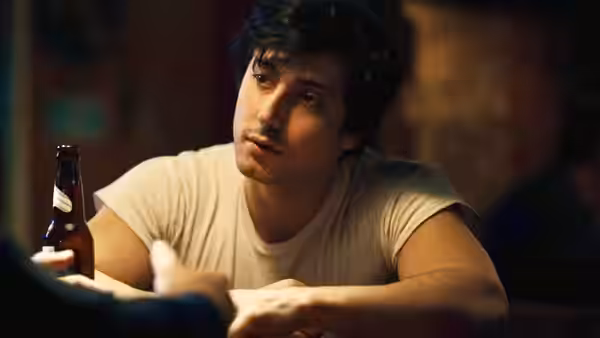 A young man sits at a bar holding a beer bottle and looking aside
