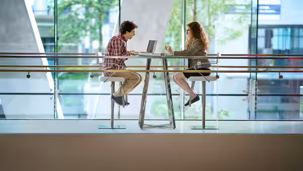 A man and woman sit across a laptop at a high table in a glass office
