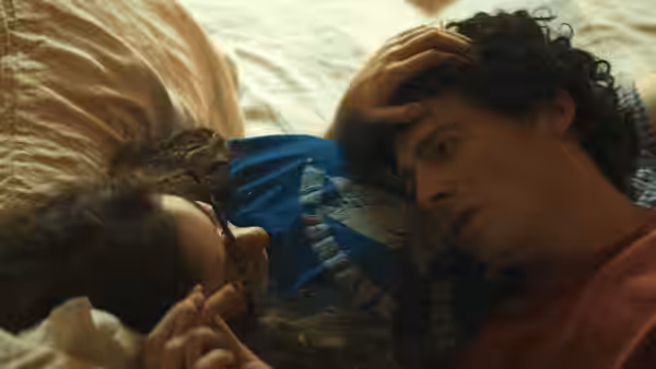 A curly-haired man lies beside a woman on a bed