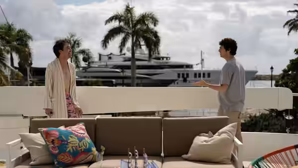 Two men talk beside a rooftop lounge overlooking palm trees and a yacht