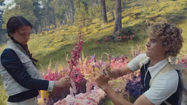 A curly-haired young man reaches toward a bloodied girl among wildflowers
