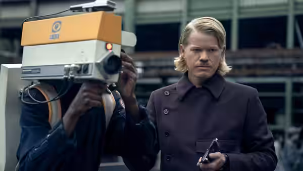 A blond man holds a small device beside a person whose face is hidden by a camera