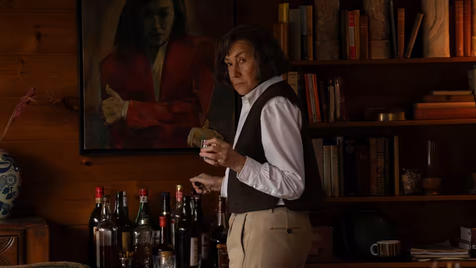 Patricia Highsmith (Helen Mirren) stands in a wood-paneled library, holding a drink