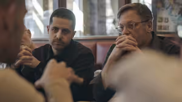Two men in conversation at a restaurant table one with glasses resting his chin on his hands