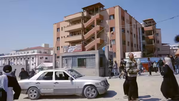 People gathered around a silver sedan on a dusty street near apartment buildings