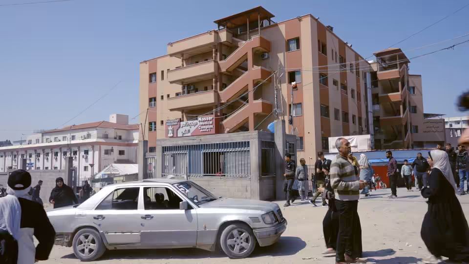 People gathered around a silver sedan on a dusty street near apartment buildings