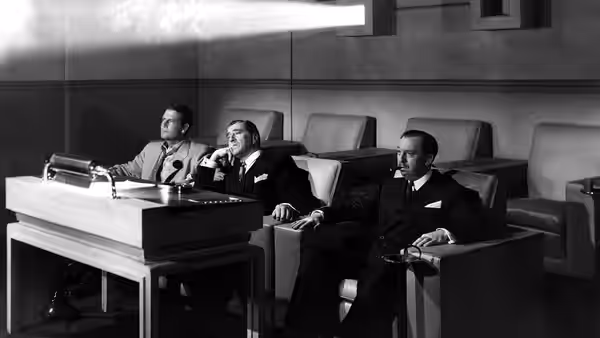 Three men in suits sit around desk with typewriter in office