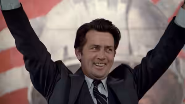 Greg Stillson (Martin Sheen) smiling with both arms raised in the air
