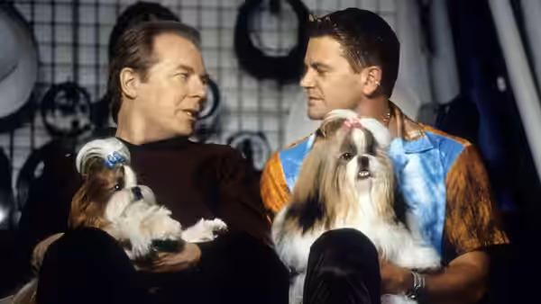 Two men hold small dogs while seated before a wall of ribbons